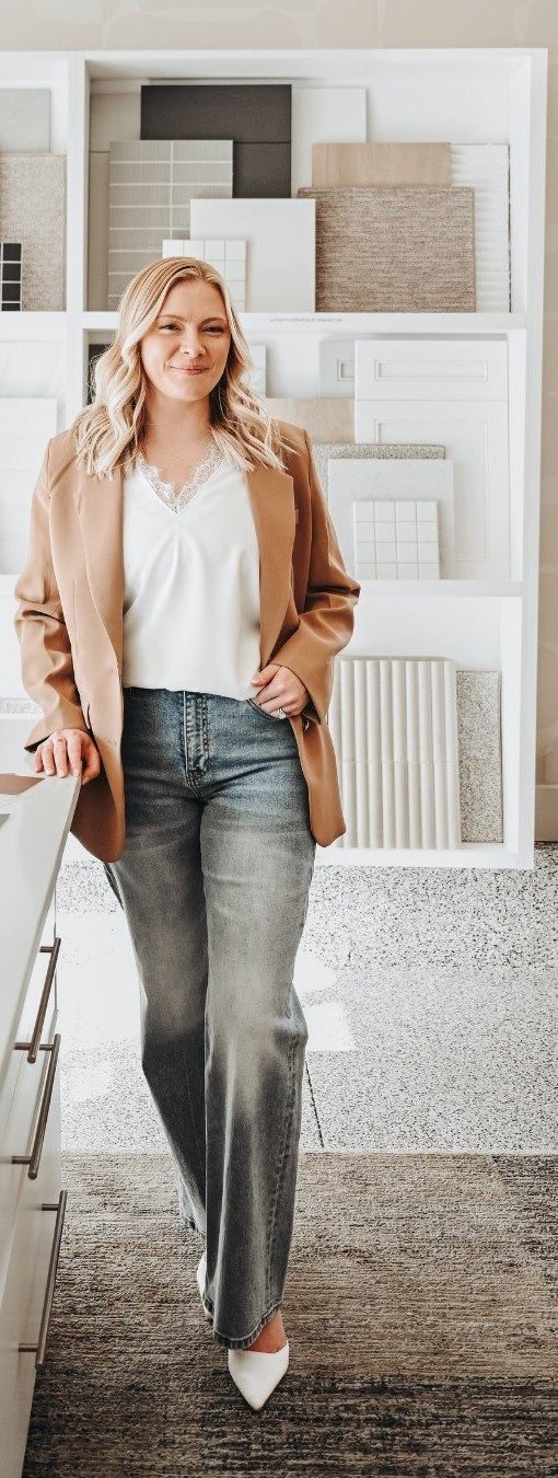 Kyrsten Smith smiling, standing in a bright room lined with design samples, wearing a camel blazer and jeans.
